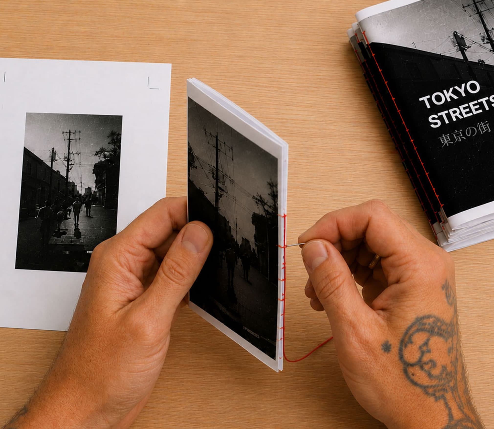 A pair of hands stitching a folded zine with red thread beside the printed sheet it was trimmed from, with a stack of finished Tokyo Streets photo zines alongside.